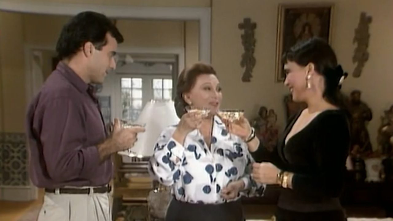 Três pessoas em um ambiente interno, levantando taças em um brinde. A mulher no centro veste uma blusa branca com estampa de flores azuis, enquanto as duas outras pessoas, à esquerda e à direita, usam roupas casuais e sorriem. O ambiente é decorado com itens de decoração clássicos e peças religiosas.