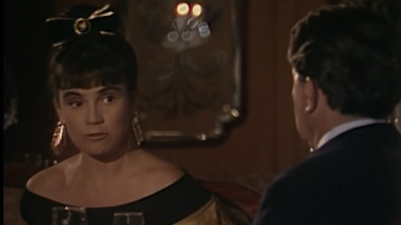 Cena mostra uma mulher e um homem sentados em uma mesa de restaurante elegante, com iluminação baixa e taças à frente. A mulher usa vestido preto de ombros à mostra, brincos longos e um laço no cabelo, enquanto fala com o homem à sua frente