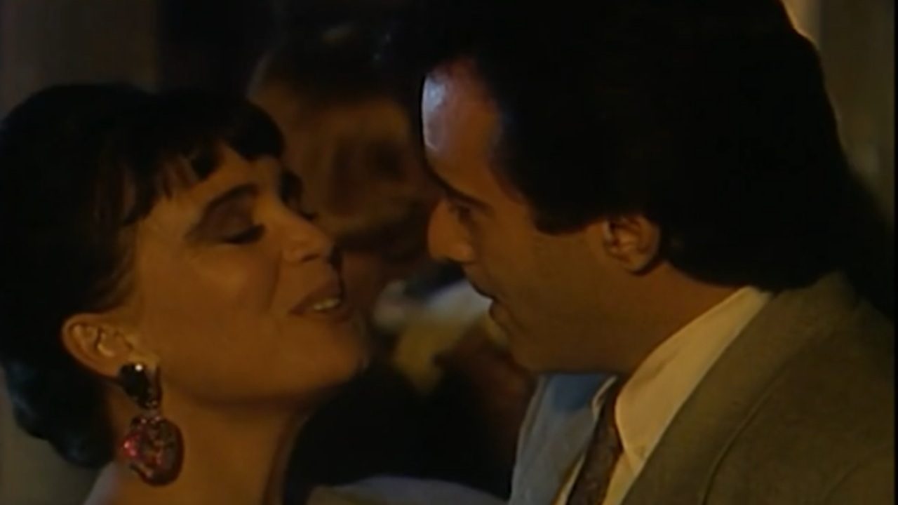 Mulher com brincos grandes sorri de olhos fechados enquanto conversa de perto com homem de terno em ambiente noturno