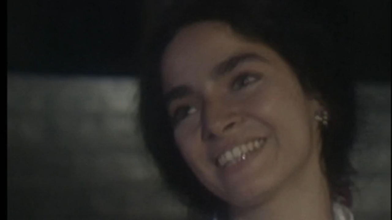 A imagem mostra uma mulher sorrindo levemente em um ambiente com pouca iluminação. Ela tem cabelos curtos e escuros, usa brincos delicados e parece olhar para alguém fora de quadro, transmitindo uma expressão de ternura e tranquilidade