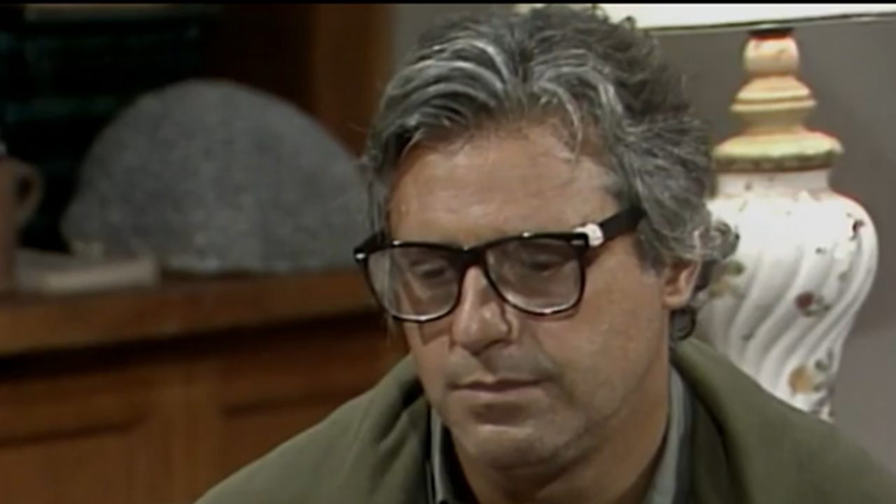 Homem de meia-idade usa óculos de armação grossa e expressão séria enquanto olha para baixo, sentado em um ambiente interno com móveis e uma luminária ao fundo