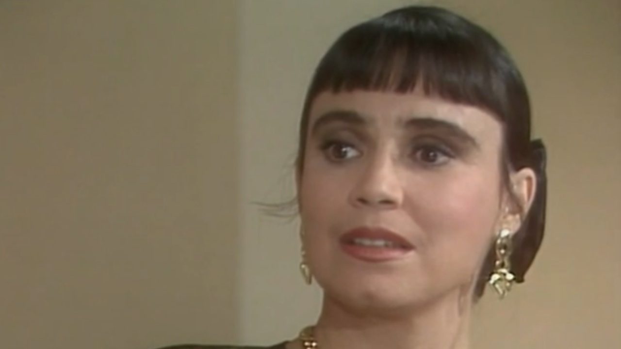 Mulher aparece em close, em ambiente interno de tom neutro, com expressão de surpresa ou apreensão, olhos bem abertos e boca levemente entreaberta. Ela tem cabelo escuro preso para trás, com franja reta curta, usa brincos dourados e colar delicado. A maquiagem é discreta, destacando os olhos. O enquadramento fechado e a expressão facial sugerem uma cena dramática, possivelmente de novela ou produção televisiva de época.