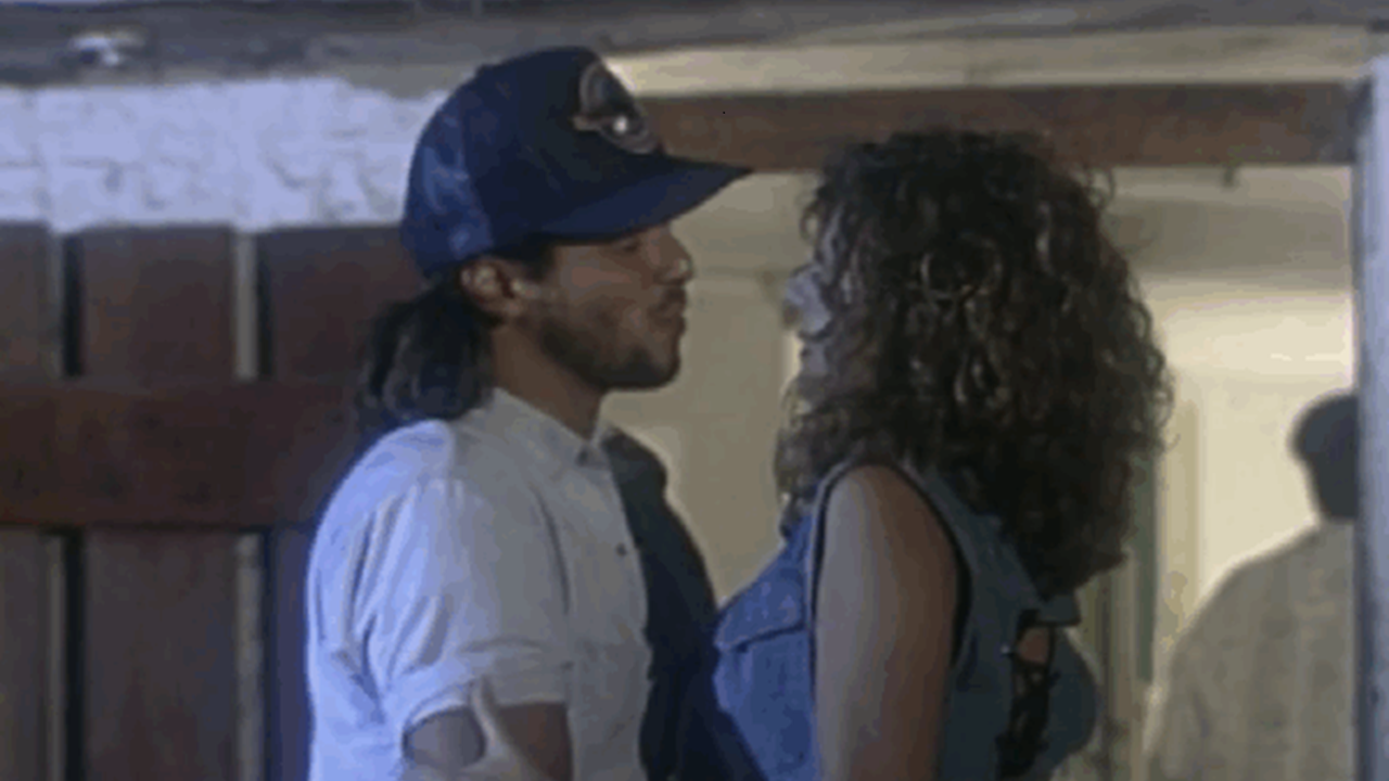 A imagem mostra um homem e uma mulher próximos um do outro, em um ambiente interno com iluminação suave. O homem usa boné azul e camisa clara, enquanto a mulher tem cabelos cacheados e veste uma blusa jeans sem mangas