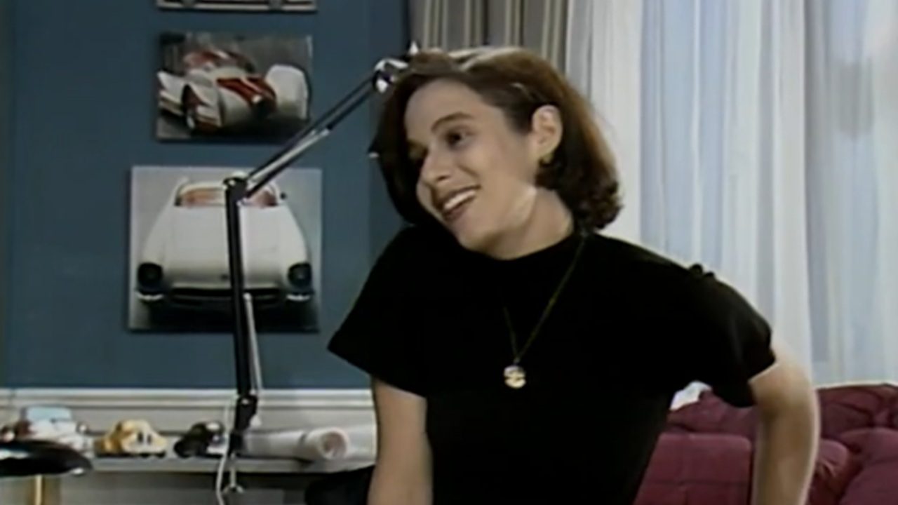 Mulher jovem, de cabelos curtos castanhos, sorri inclinando levemente a cabeça em um ambiente interno. Ela veste blusa preta e colar dourado. Ao fundo, há uma parede azul com quadros de carros esportivos, uma luminária articulada sobre a mesa e cortinas claras ao lado de um sofá vermelho.