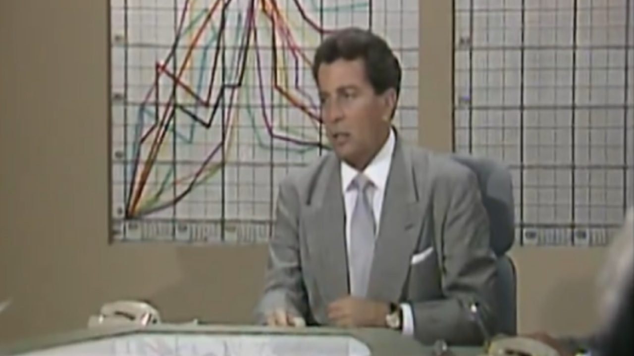 Homem de terno cinza e gravata clara está sentado atrás de uma mesa com telefones enquanto fala em ambiente de escritório com paredes bege e dois grandes gráficos coloridos com linhas ascendentes e descendentes presos na parede ao fundo