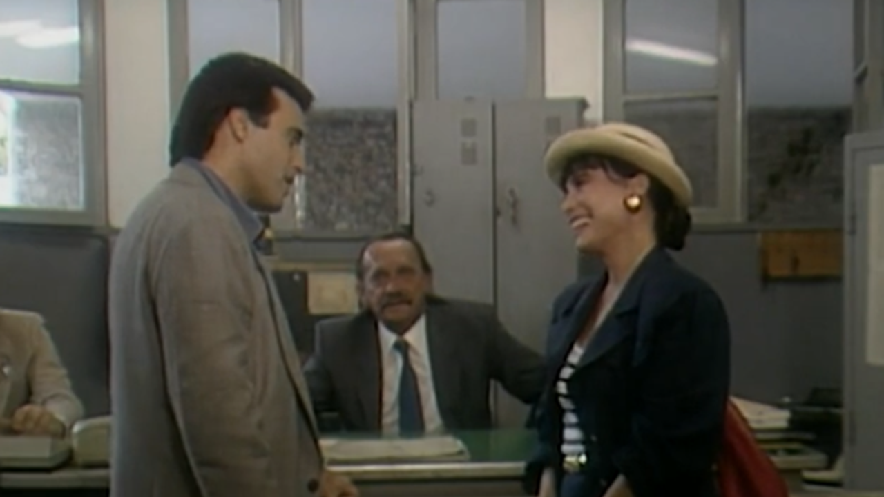 Cena mostra três pessoas em um escritório antigo: um homem e uma mulher em pé, sorrindo um para o outro, enquanto outro homem, sentado atrás de uma mesa, os observa. O ambiente tem janelas grandes, armários metálicos e iluminação típica de repartições públicas, sugerindo um clima leve e cotidiano