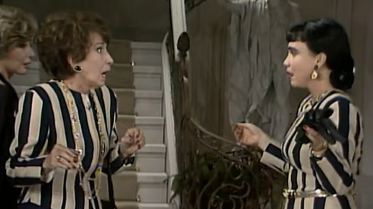 Duas mulheres em um ambiente interno discutem próximas a uma escada. Ambas usam roupas listradas em preto e branco. A mulher à esquerda demonstra surpresa, com expressão de espanto e mãos erguidas. A mulher à direita gesticula enquanto segura um objeto escuro. Ao fundo, vê-se a escadaria e parte da decoração da casa.