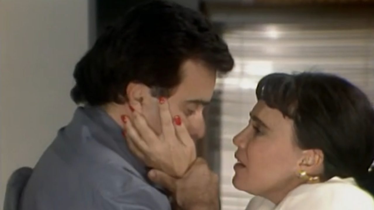 Cena mostra um homem e uma mulher em momento emocional e íntimo. A mulher segura o rosto do homem com as duas mãos, enquanto eles conversam muito próximos um do outro. A iluminação suave e o enquadramento fechado destacam a tensão e a vulnerabilidade da interação