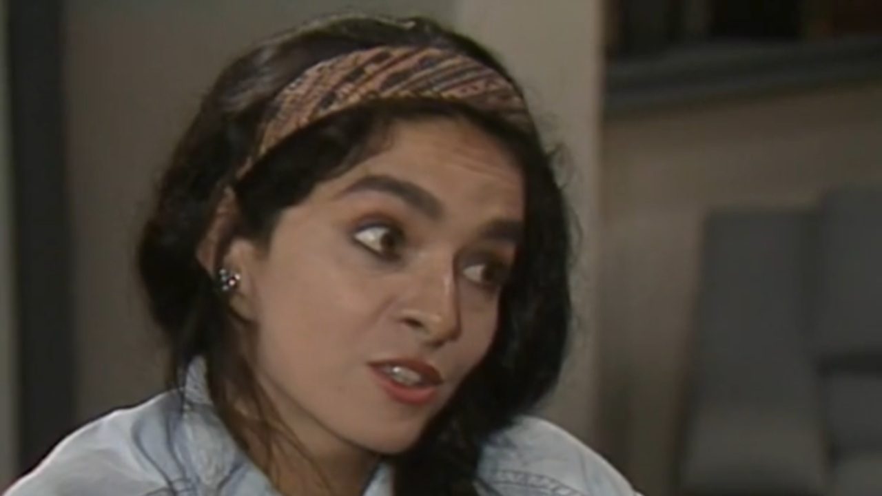 Mulher de cabelos escuros e levemente ondulados, usando uma faixa estampada na cabeça e camisa clara, olha para o lado com expressão desconfiada em um ambiente interno.