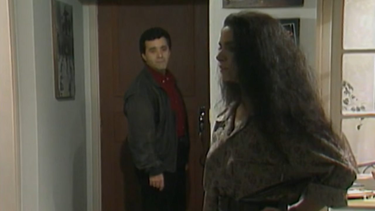 Cena em ambiente interno mostra um homem parado à porta de um apartamento, olhando para dentro, enquanto uma mulher aparece em primeiro plano, de perfil, com a mão apoiada na cintura. O homem veste camisa vermelha e jaqueta escura; a mulher usa blusa de mangas longas e tem cabelos longos e soltos. O espaço é iluminado por luz natural que entra por uma janela à direita, sugerindo um momento de tensão ou despedida dentro de um ambiente doméstico.