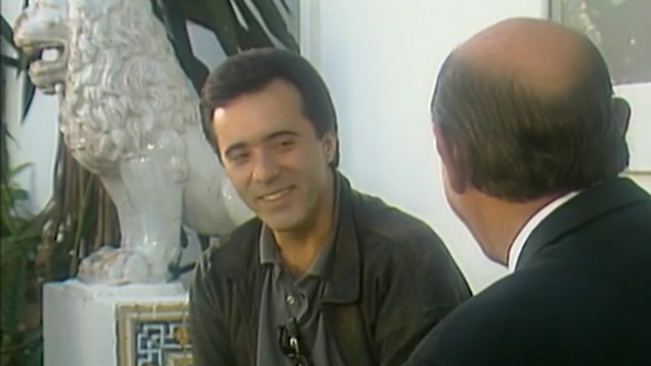 Tony Ramos em cena da novela Rainha da Sucata conversa com outro personagem em ambiente interno com escultura decorativa ao fundo