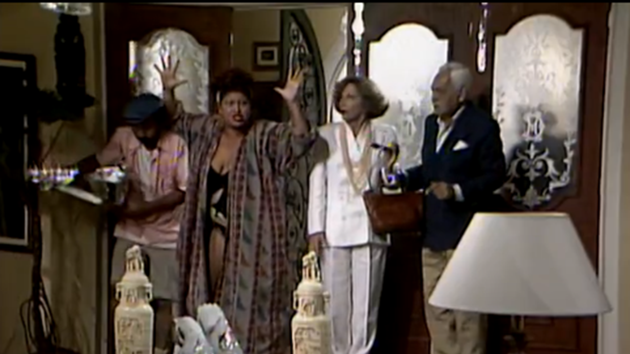 A imagem mostra uma cena de comédia em um ambiente doméstico, com quatro pessoas próximas à porta de entrada. Uma mulher ao centro parece muito exaltada, gesticulando com as mãos levantadas, enquanto os outros três observam com surpresa ou preocupação. O cenário tem decoração clássica, com vasos e móveis elegantes.