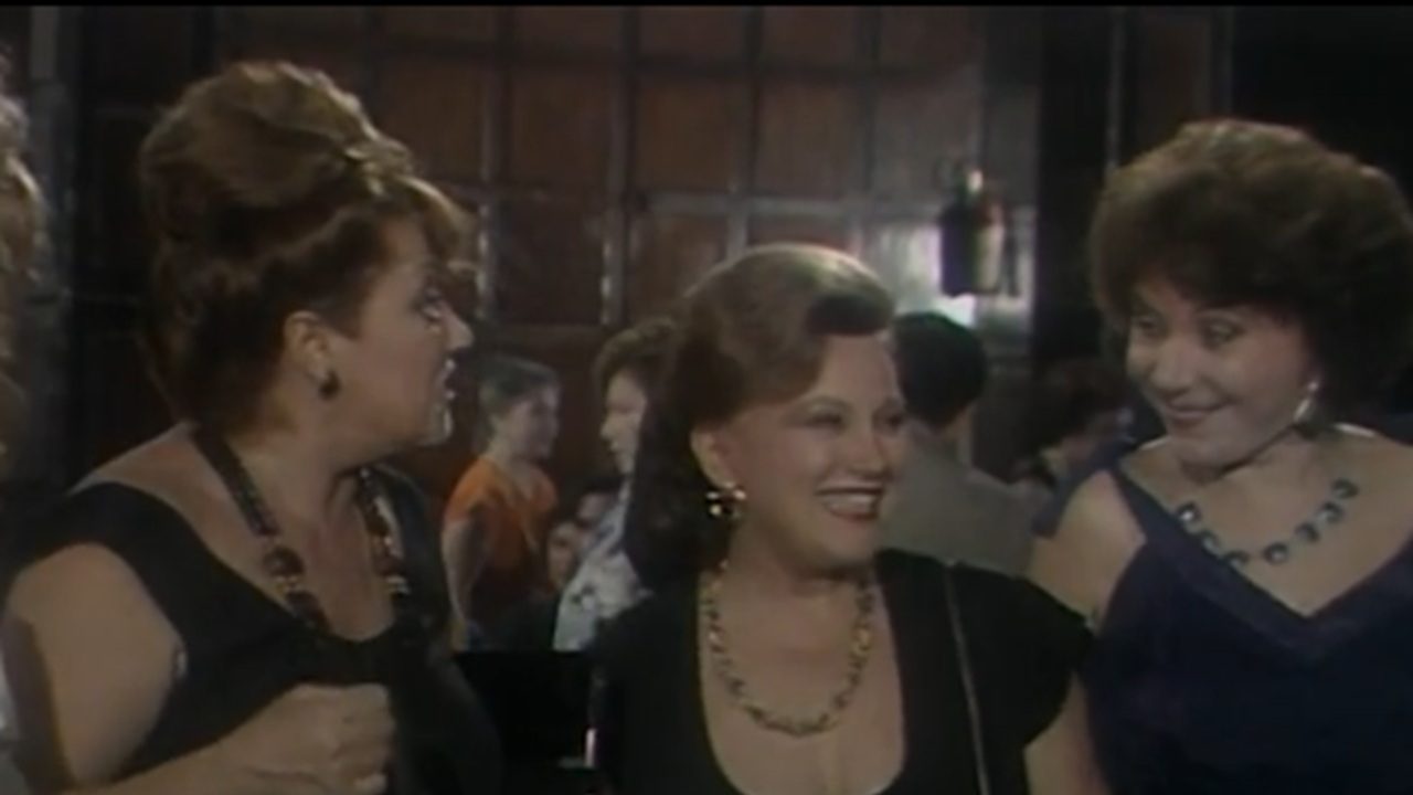 Três mulheres vestidas para uma ocasião social conversam e sorriem em um ambiente interno movimentado, com outras pessoas ao fundo. Elas usam vestidos elegantes e joias chamativas, e a iluminação suave destaca o clima descontraído do encontro.