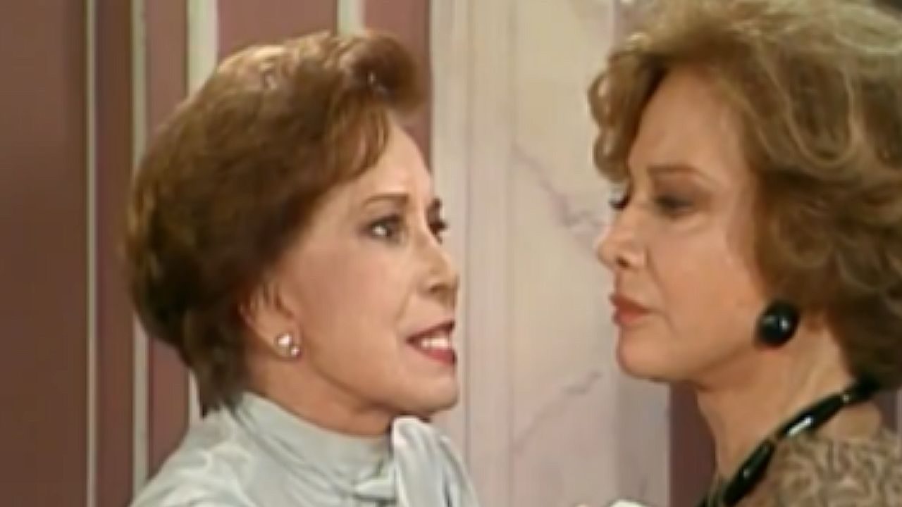 Duas mulheres discutem frente a frente com expressões tensas em cena de novela em ambiente interno