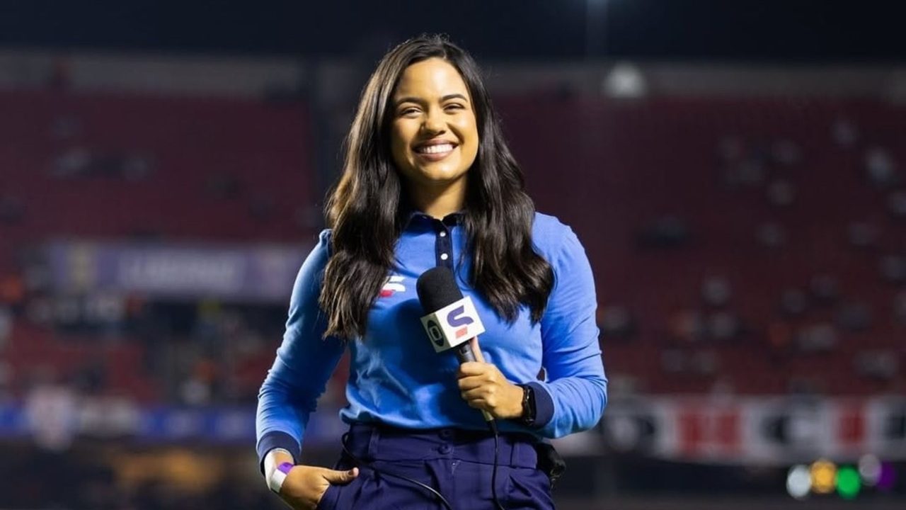 Uma repórter esportiva sorri para a câmera enquanto segura um microfone em um estádio de futebol à noite. Ela veste uma blusa azul de manga longa e calça escura, e há arquibancadas vermelhas desfocadas ao fundo