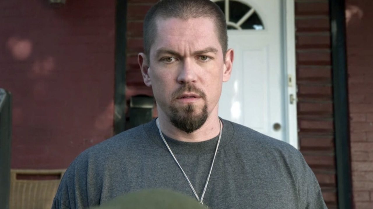 Homem de expressão séria com barba rala e camiseta cinza aparece em frente a uma casa de tijolos vermelhos