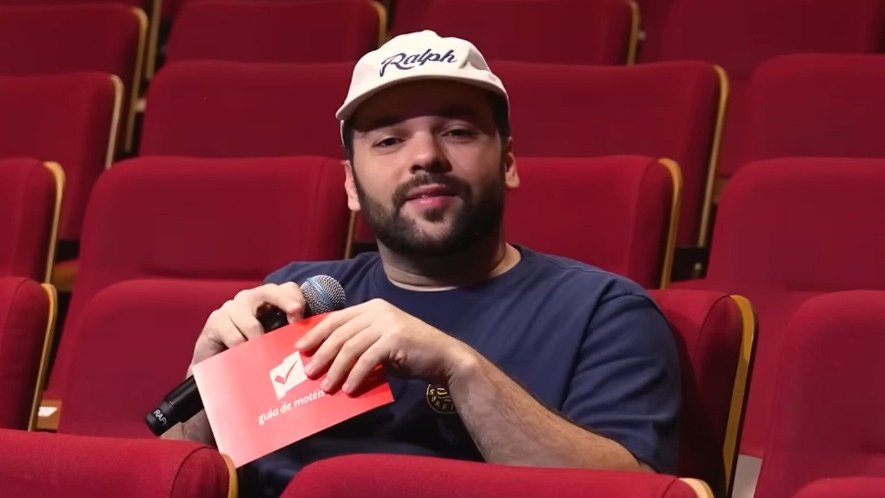 Raphael Ghanem segura microfone e cartão vermelho enquanto fala sentado em plateia de teatro com poltronas vermelhas
