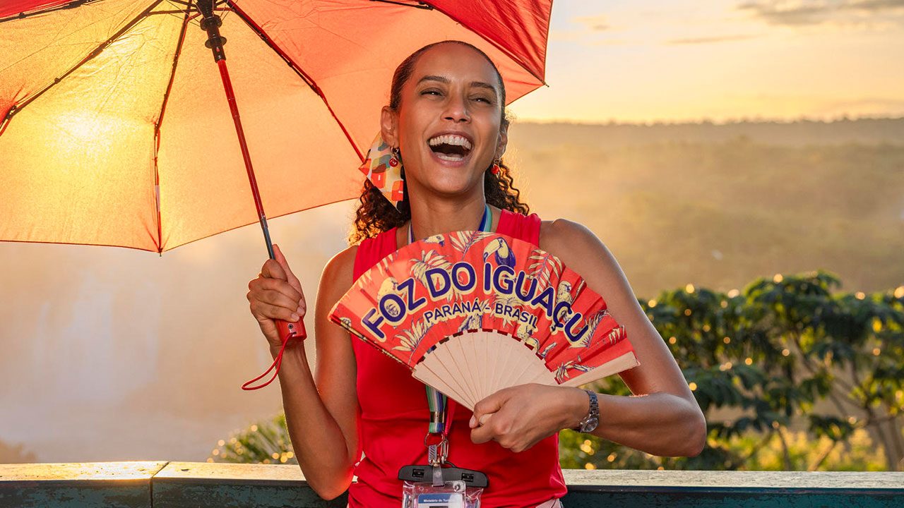 Tais Araujo caracterizada como Raquel em Vale Tudo sorrindo com sombrinha vermelha e leque escrito Foz do Iguaçu