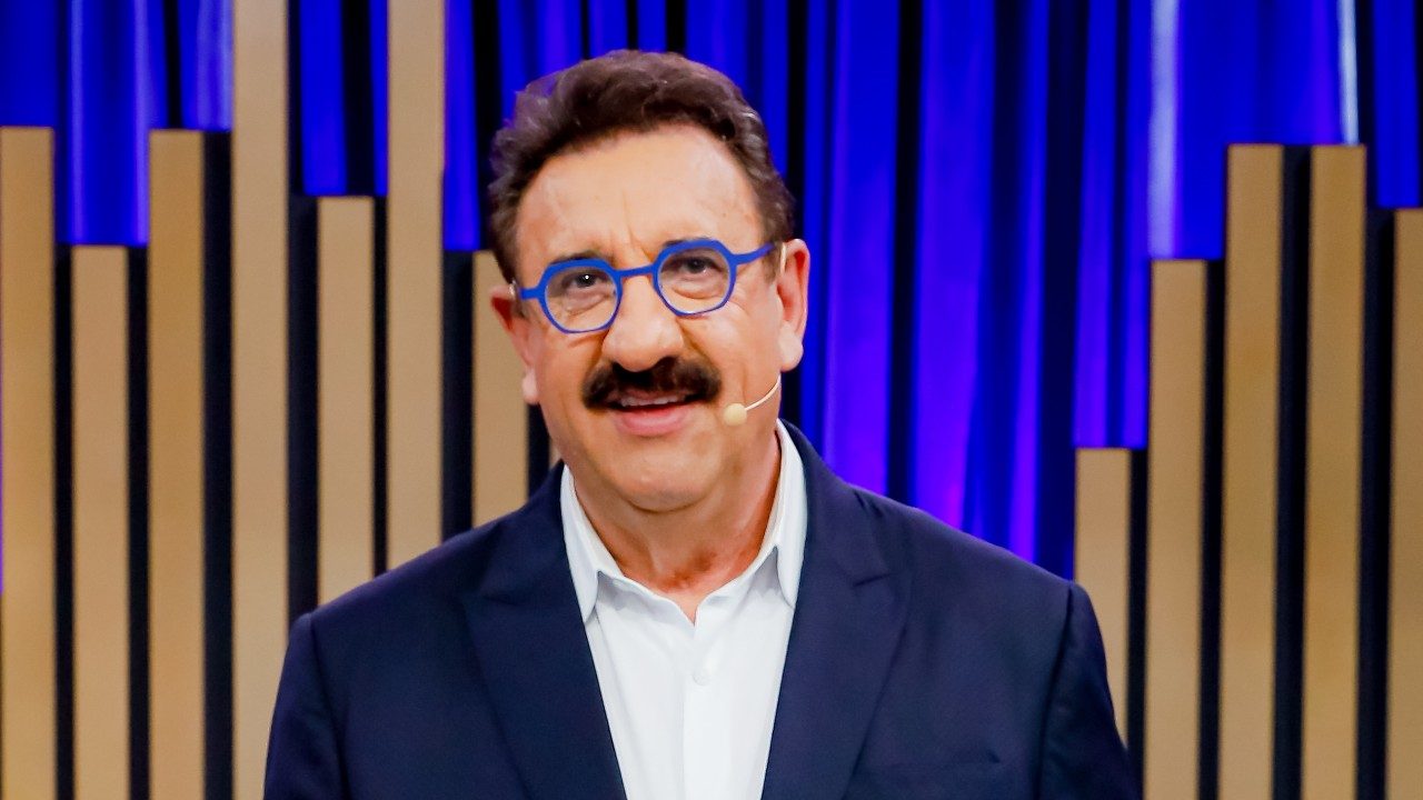 Apresentador Ratinho sorridente usa óculos de armação azul, bigode marcante e paletó azul escuro sobre camisa branca, no cenário do Programa do Ratinho