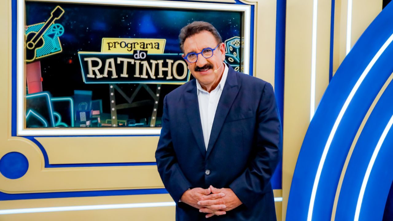 Apresentador Ratinho sorri de terno azul e óculos de armação colorida, posicionado à frente do telão com a logomarca de seu programa no cenário do SBT