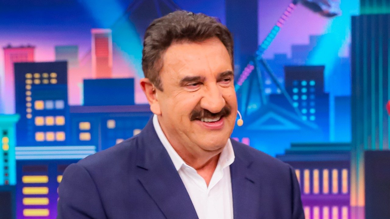 Ratinho sorri com microfone de lapela durante gravação de programa em estúdio com cenário colorido de cidade ao fundo