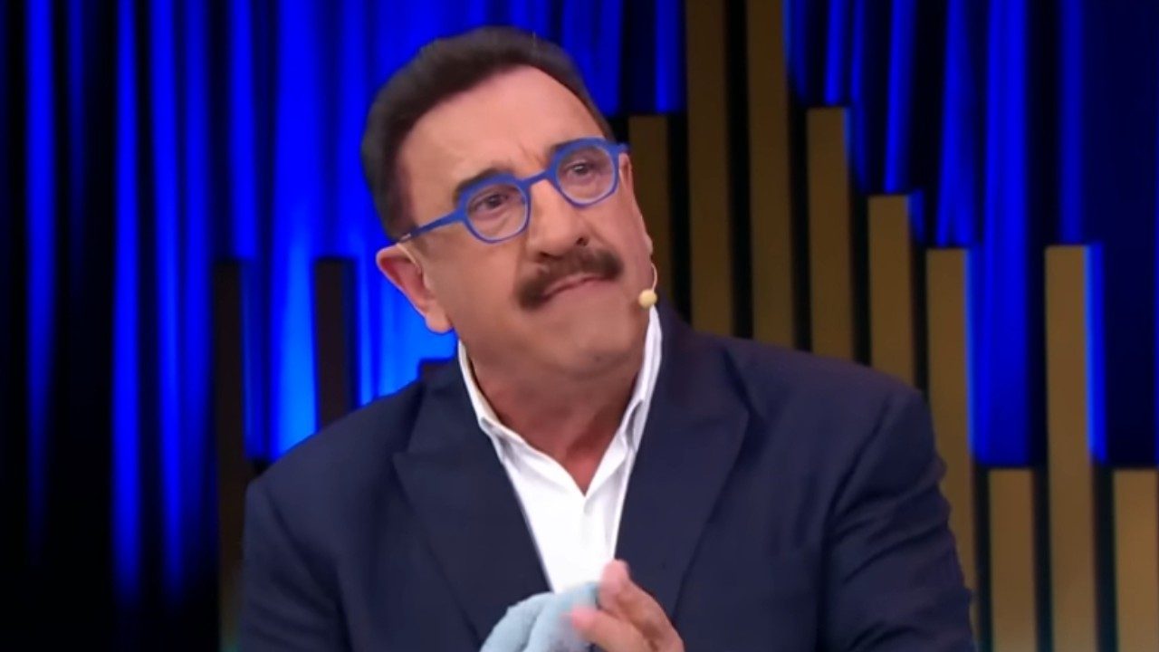 Ratinho usando óculos azuis durante apresentação em estúdio com cenário iluminado ao fundo