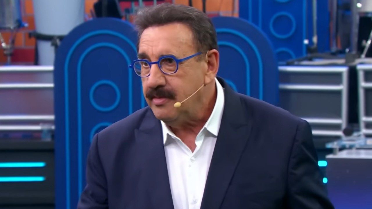 Ratinho de terno escuro e óculos azuis fala ao microfone de rosto em estúdio do Programa do Ratinho no SBT
