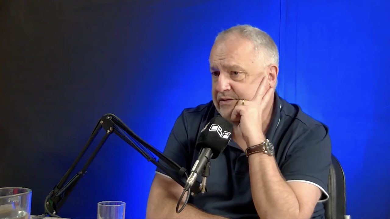 Raul Costa Jr., de camisa polo azul, apoia o rosto na mão diante de microfone em estúdio de podcast