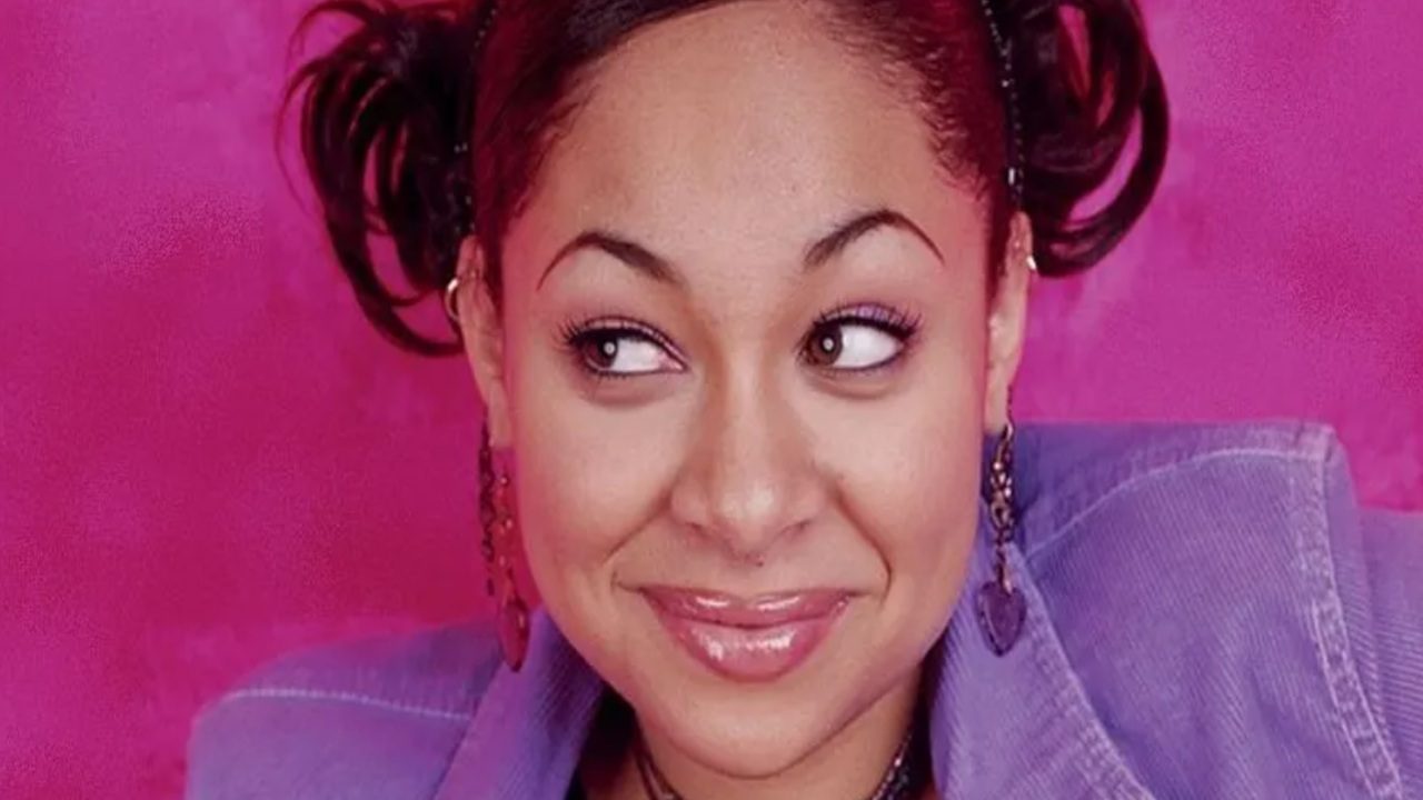 Atriz de As Visões da Raven sorri com expressão divertida, olhando para o lado, em frente a um fundo rosa vibrante. Ela usa jaqueta roxa, brincos em formato de coração e penteado com coques laterais