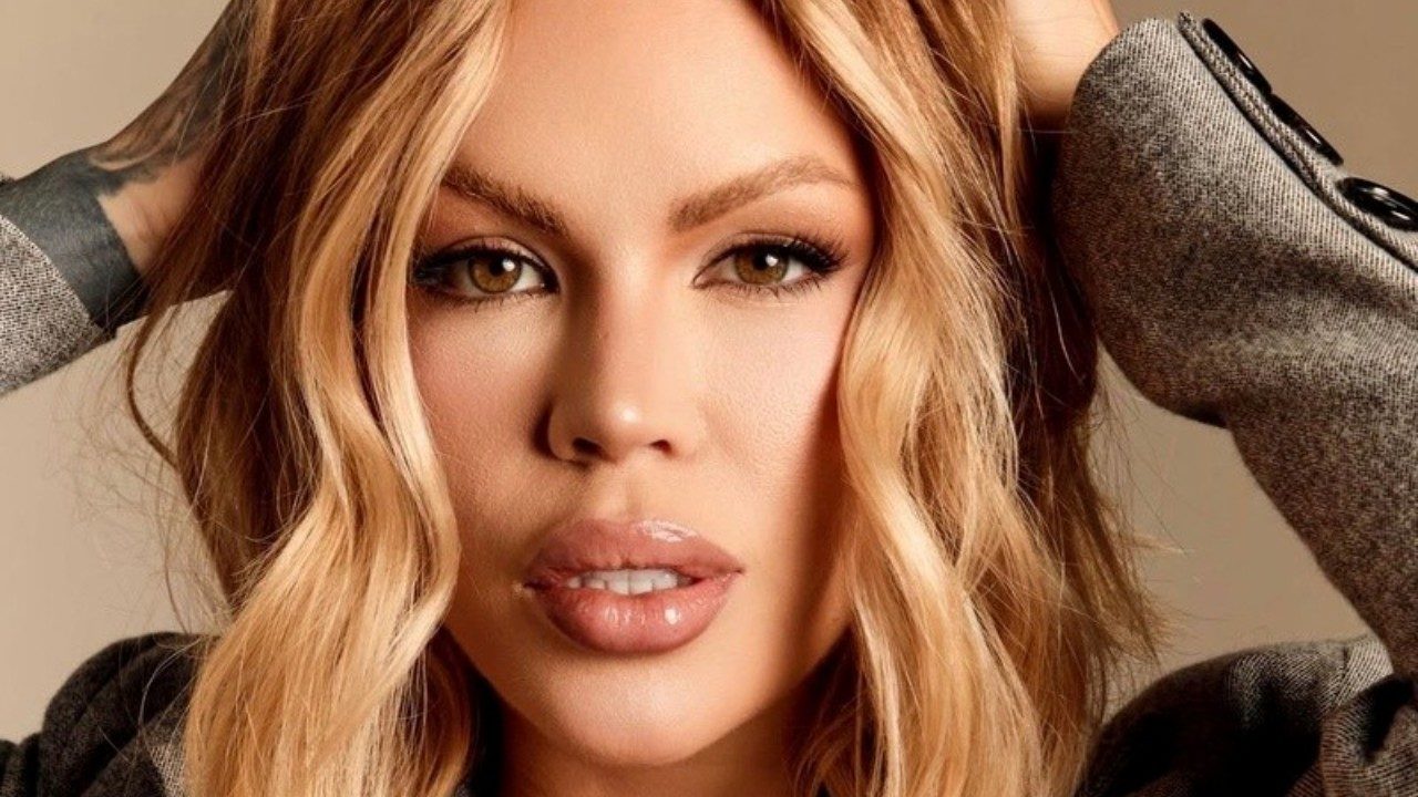 Ravena Hanniely em close com cabelo loiro ondulado mãos na cabeça e olhar direto para a câmera em ensaio fotográfico