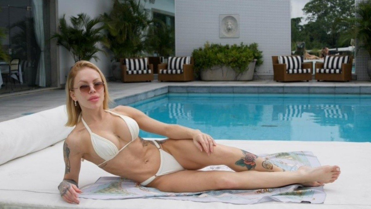 Ravena Hanniely de biquíni branco posa deitada à beira da piscina com óculos escuros em área externa com espreguiçadeiras