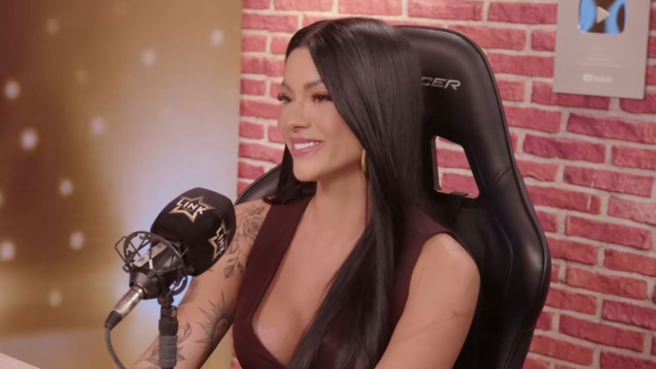 Rayane Figliuzzi sorrindo durante participação em podcast com microfone à frente e parede de tijolos ao fundo