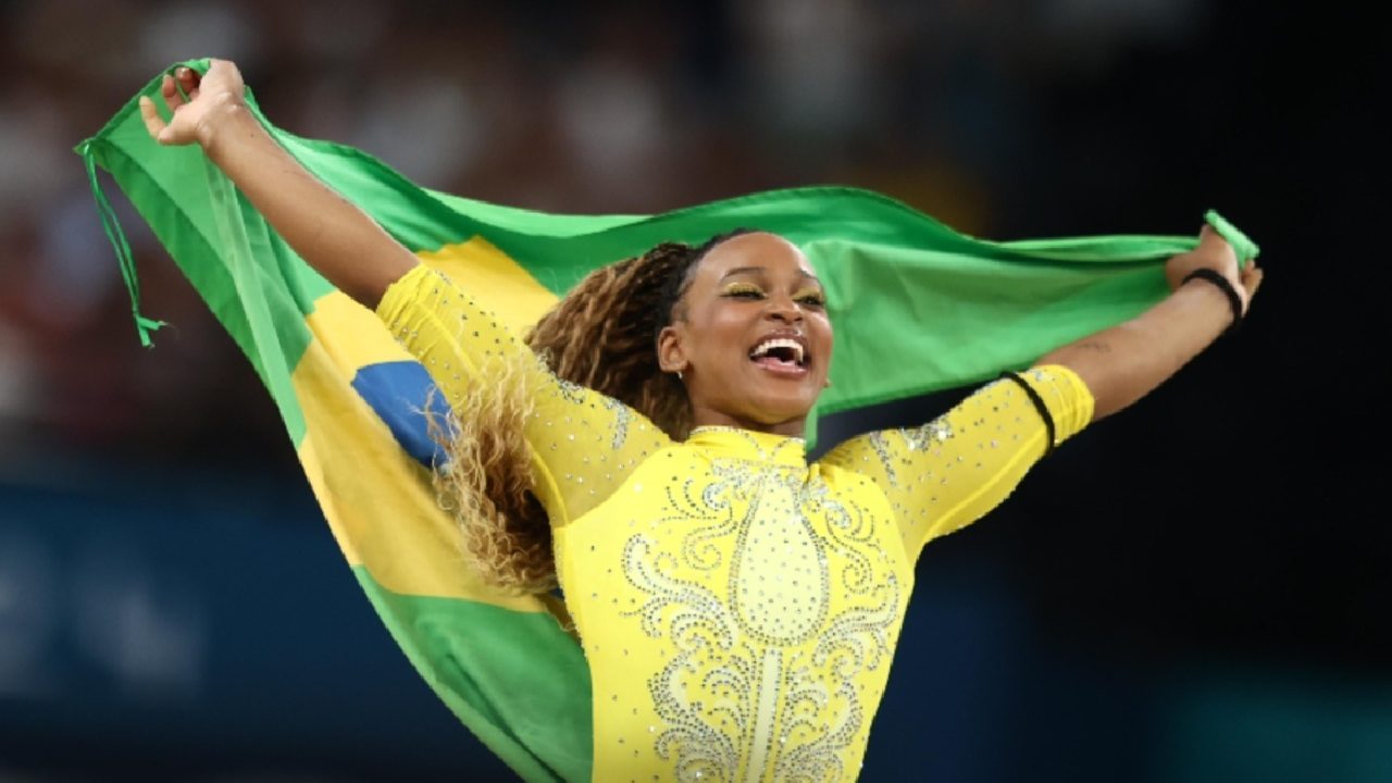 Atleta sorri e comemora segurando a bandeira do Brasil aberta sobre os ombros, vestindo uniforme amarelo com detalhes brilhantes, em uma arena esportiva lotada