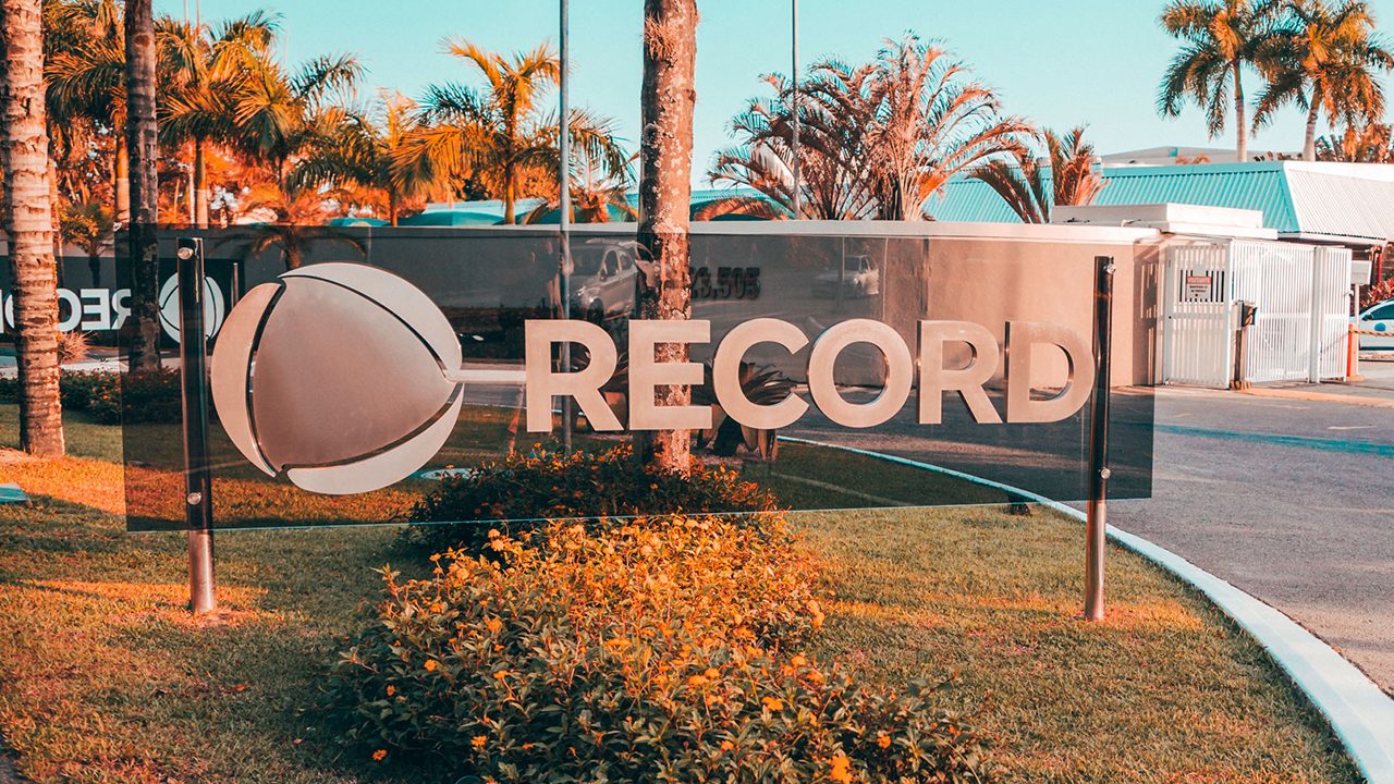 Placa com logotipo da Record na entrada da sede da emissora no Rio de Janeiro com palmeiras e guarita ao fundo