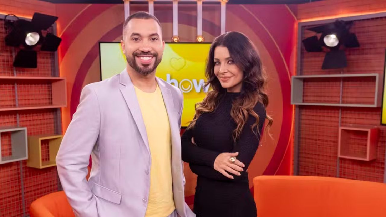 Dois apresentadores posam sorrindo em um estúdio colorido, com paredes em tons de laranja e amarelo e iluminação de TV ao fundo. O homem veste blazer claro e camiseta amarela, enquanto a mulher usa um vestido preto de mangas longas. Ambos estão de pé diante de um painel com o logo do programa