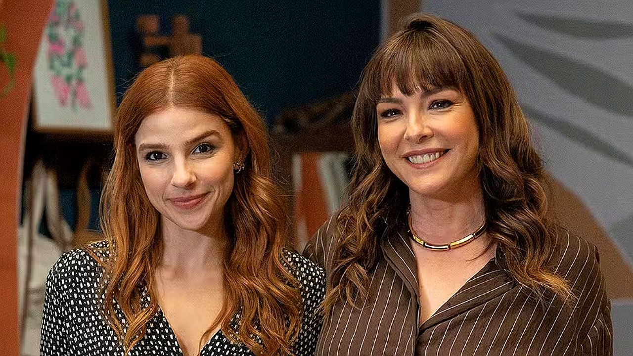 Duas mulheres sorriem lado a lado em ambiente interno com prateleiras ao fundo em cena de série ou novela