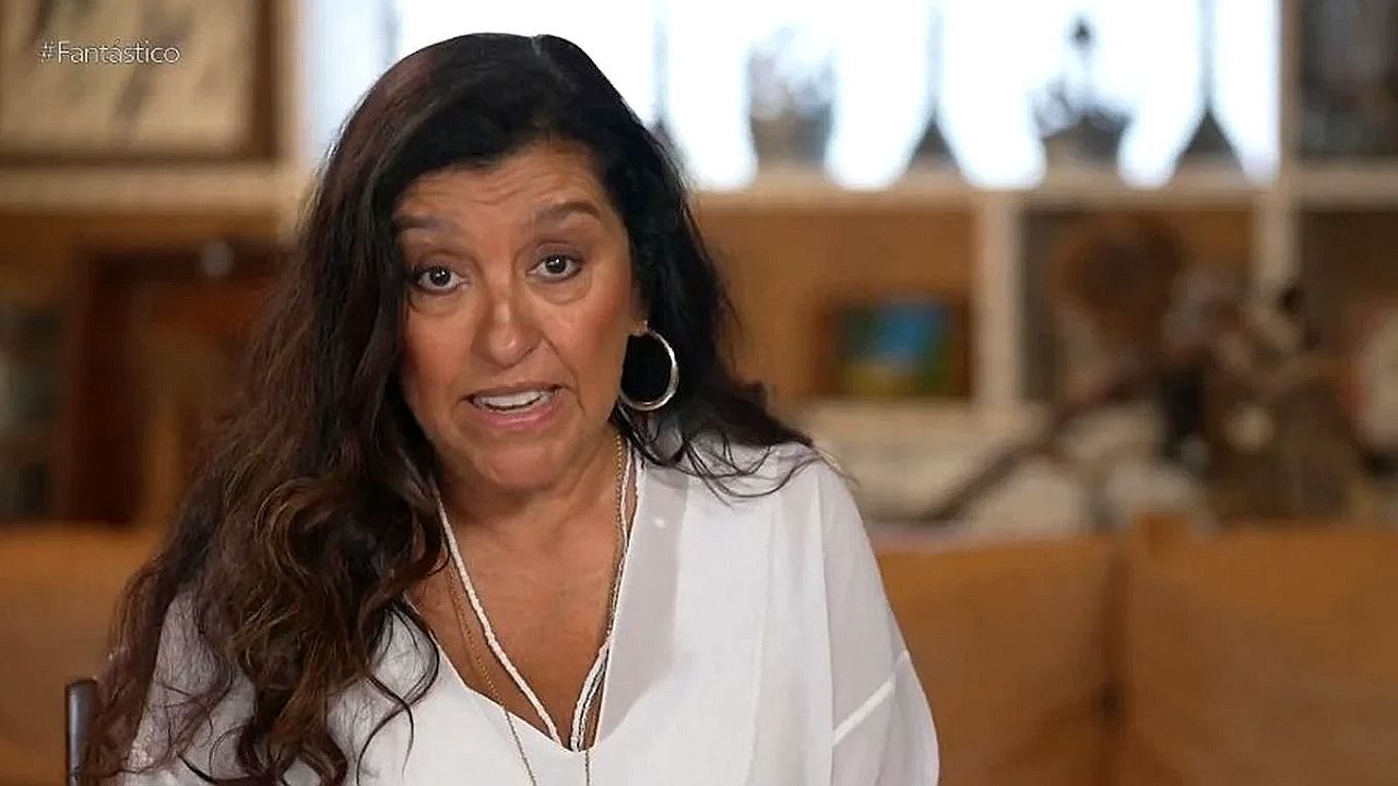 Regina Casé fala durante entrevista ao Fantástico usando blusa branca e brincos de argola