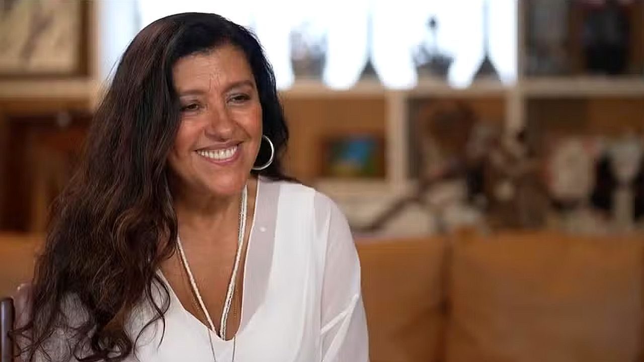 Regina Casé sorri durante entrevista em ambiente residencial usando blusa branca e acessórios prateados com estante desfocada ao fundo