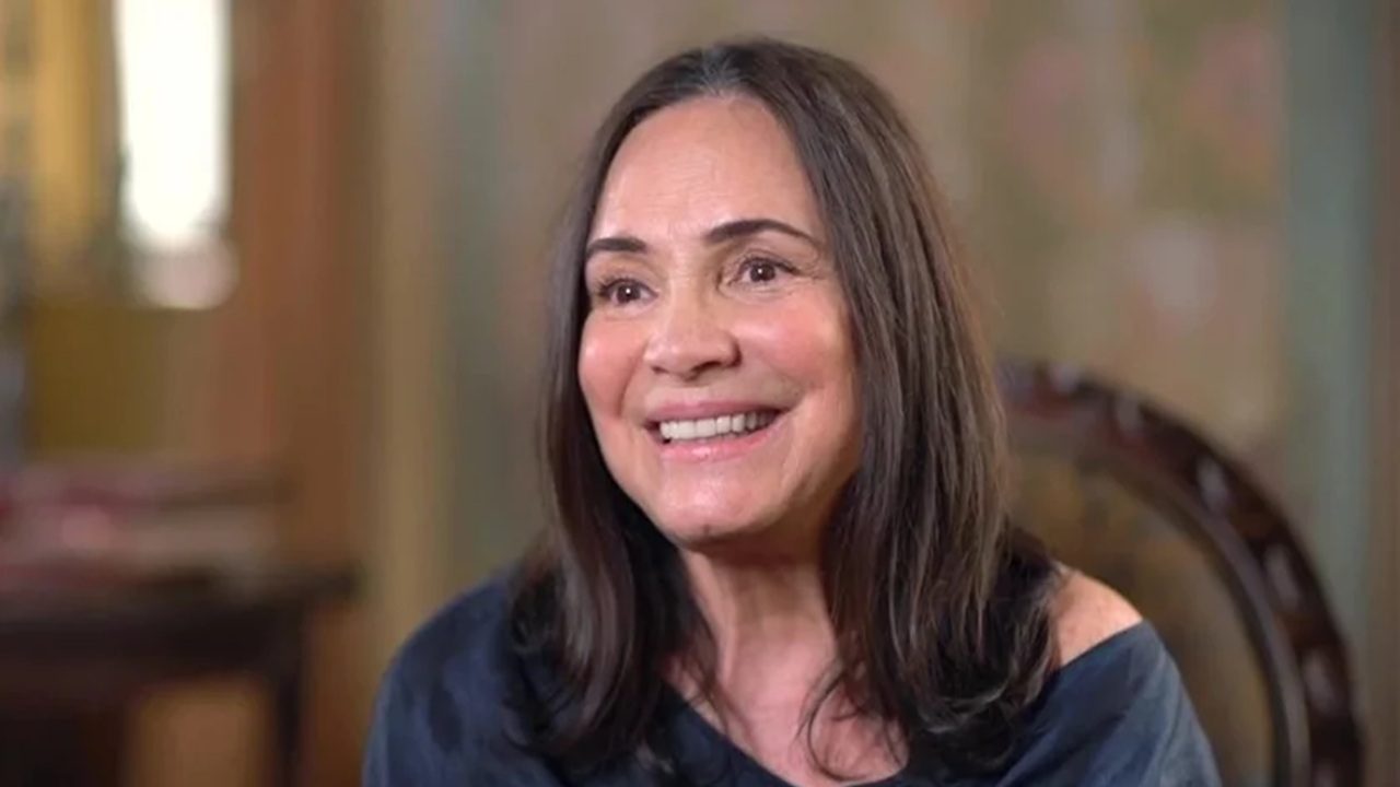 Mulher de cabelos castanhos escuros, lisos e na altura dos ombros, sorri amplamente. Ela veste uma blusa azul escura e está em um ambiente interno com decoração desfocada ao fundo, transmitindo sensação de entrevista ou conversa descontraída