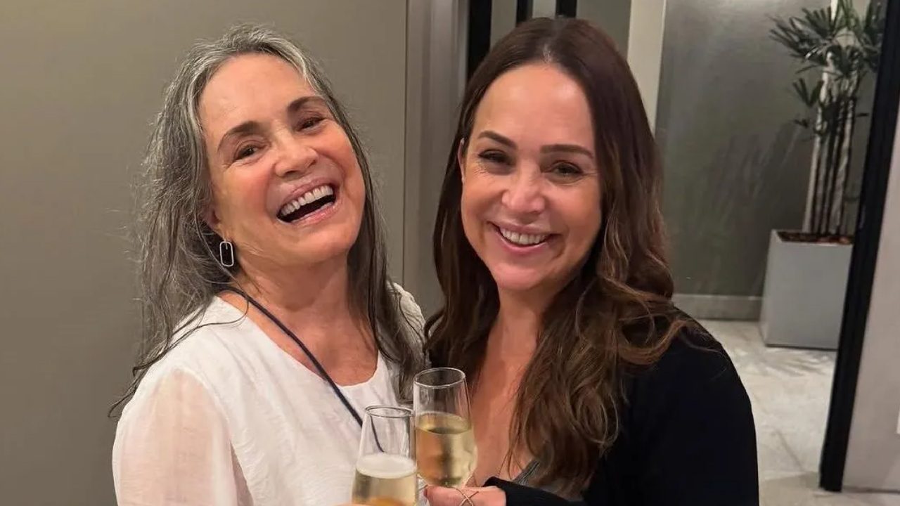 Regina Duarte e Gabriela Duarte sorrindo lado a lado, brindando com taças de espumante em ambiente interno iluminado, em clima de celebração e descontração