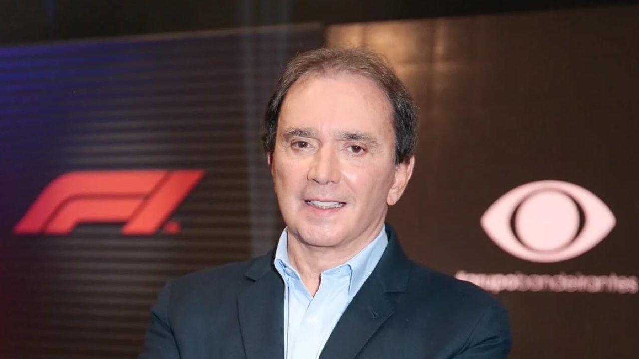 Homem de terno posa sorridente em frente a painéis com os logotipos da Fórmula 1 e de uma emissora de televisão, sugerindo evento ou cobertura esportiva