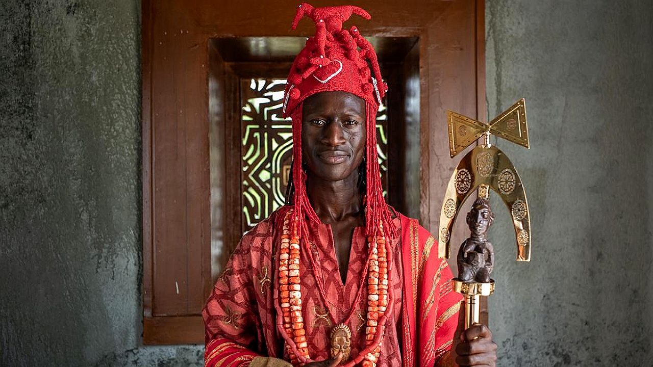 Um homem está em pé diante de uma porta de madeira, vestindo trajes tradicionais em tons de vermelho. Ele usa um adorno de cabeça elaborado, com detalhes e extensões, além de colares de contas grossas que cobrem o peito. Em uma das mãos, segura um objeto cerimonial decorado, com elementos metálicos e uma pequena escultura. Sua expressão é serena e direta. O ambiente ao redor sugere um espaço cultural ou histórico, com paredes texturizadas e elementos artesanais.