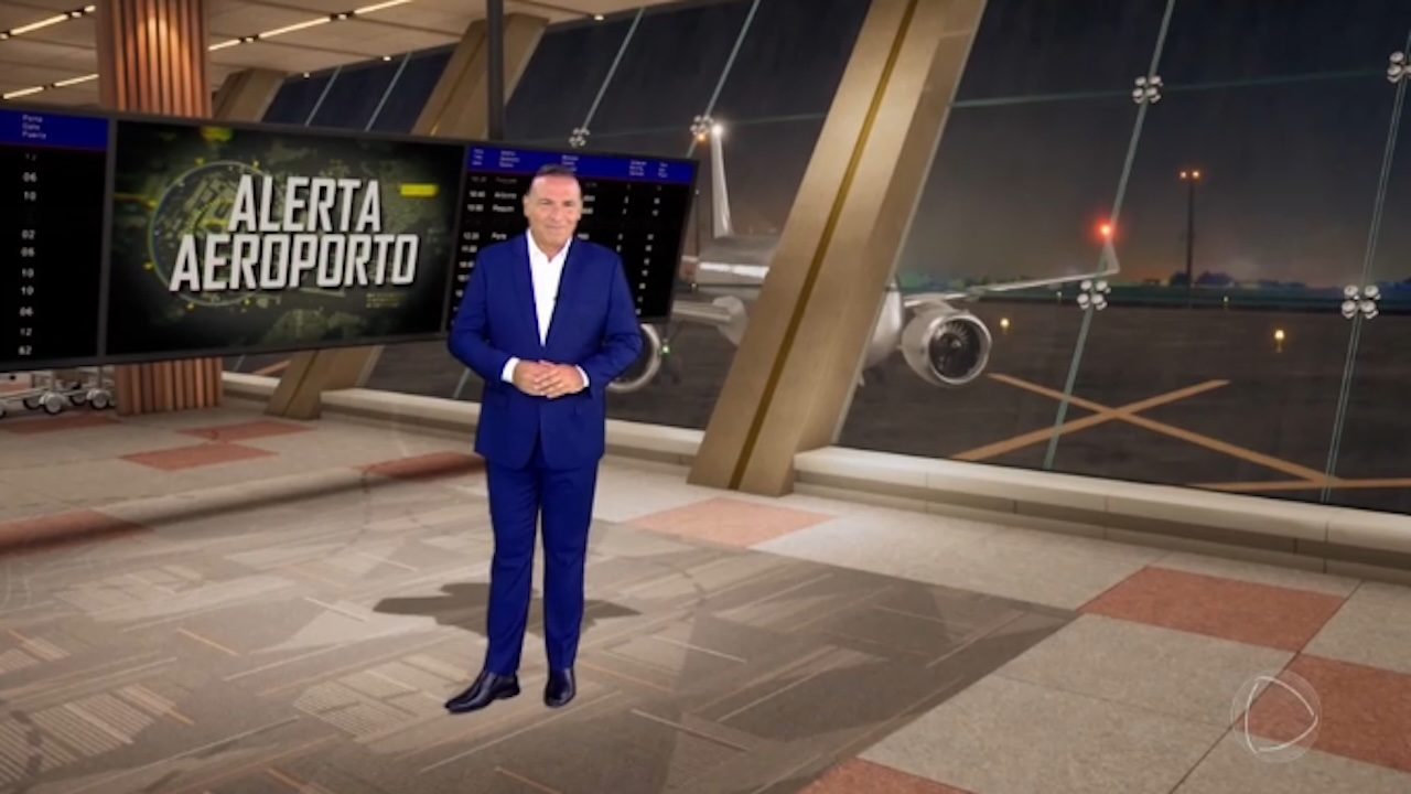 Reinaldo Gottino, de terno azul, em pé no estúdio do Alerta Aeroporto diante de telão e pista com avião ao fundo