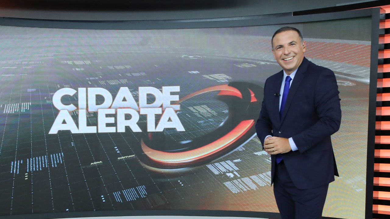 Reinaldo Gottino no estúdio do Cidade Alerta da Record diante do telão com o logotipo do telejornal