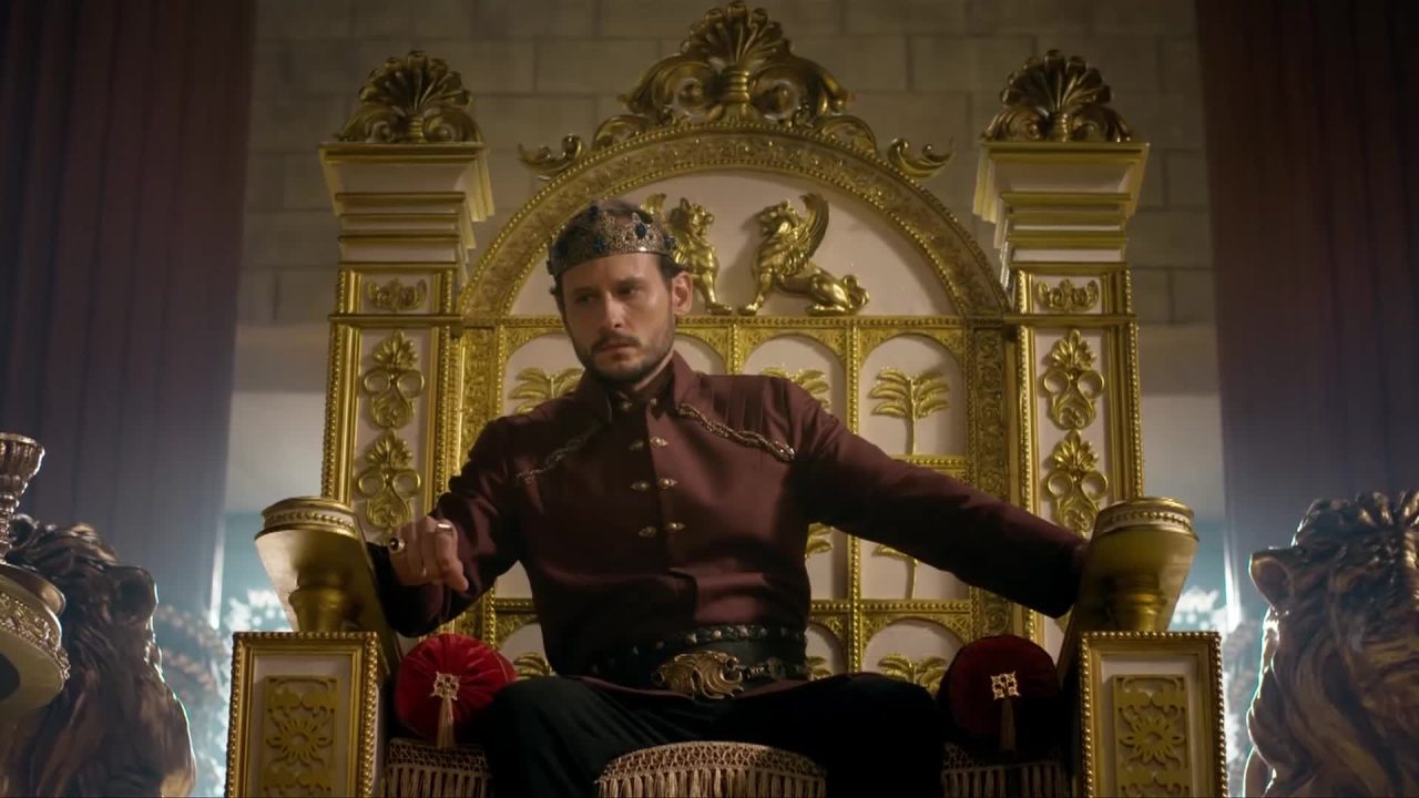 Salomão (Guilherme Dellorto) com coroa e túnica escura sentado em trono dourado ornamentado em cena da novela Reis, exibida pela Record