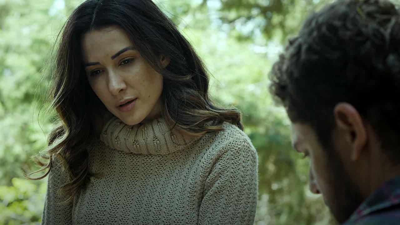 Mulher de cabelos longos e castanhos usa suéter bege de gola alta enquanto conversa com homem em área arborizada ao ar livre