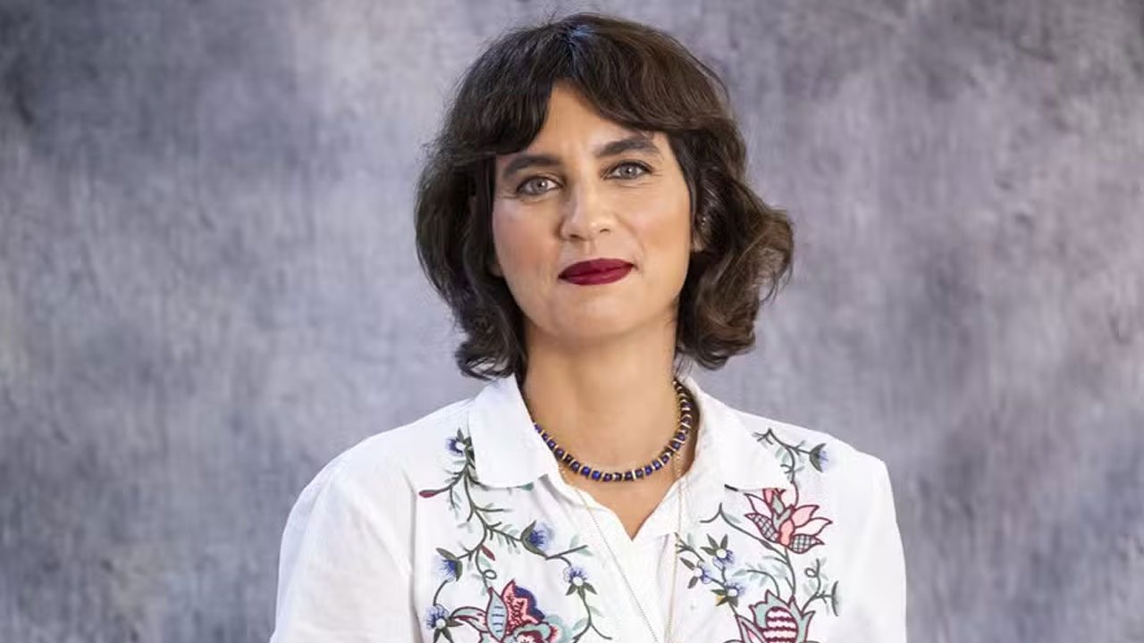 Renata Corrêa com cabelo escuro, batom vermelho e camisa branca com bordado floral colorido olha para a câmera em foto de divulgação
