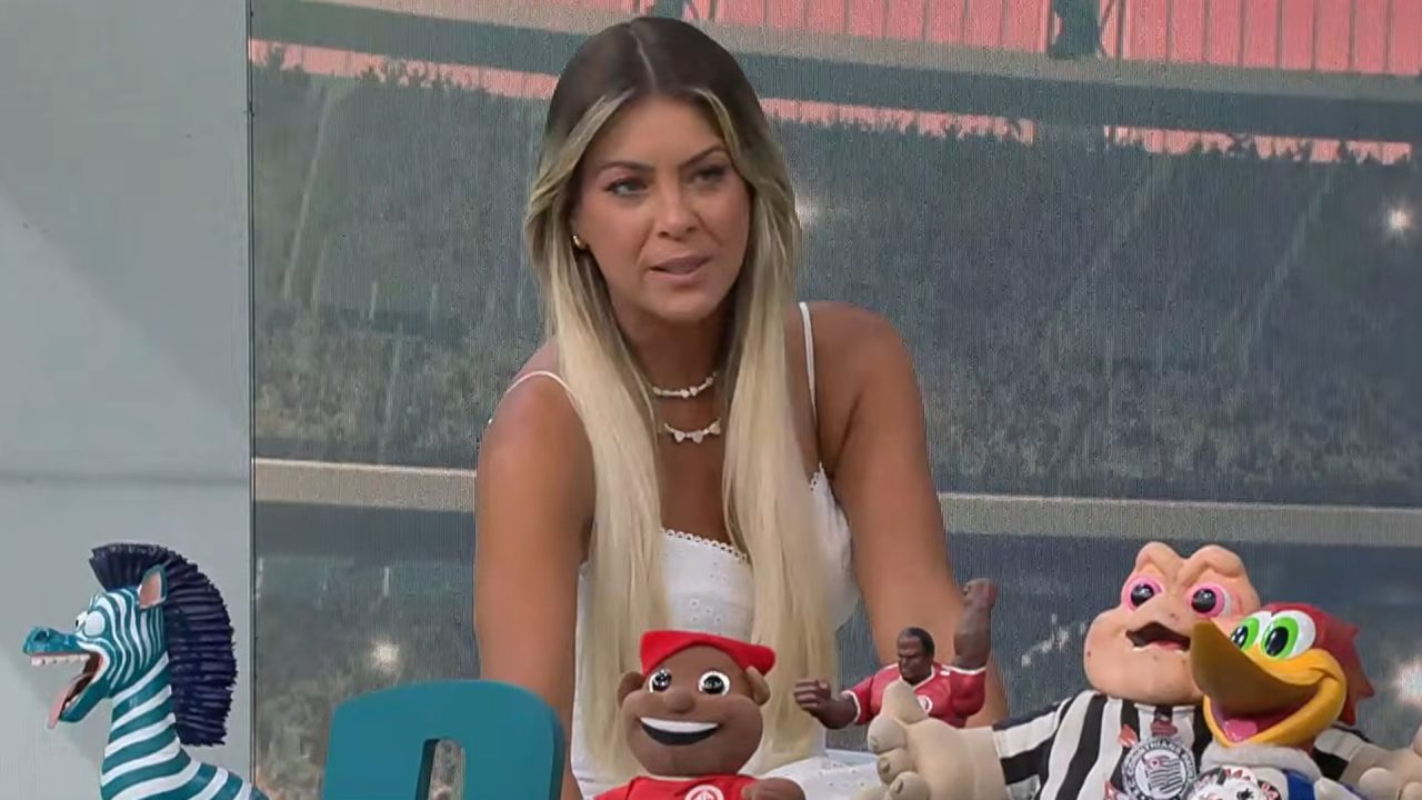 Renata Fan, apresentadora da Band, é uma mulher com cabelos longos e loiros, usando blusa branca, e está sentada à mesa do estúdio do Jogo Aberto. À sua frente há diversos bonecos e mascotes coloridos, incluindo personagens de time de futebol. Ao fundo, aparece a imagem de um estádio ao entardecer exibida em um telão.