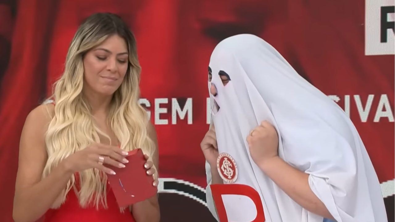 A imagem mostra uma cena bem-humorada em que Renata Fan interage com outra pessoa vestida como um fantasma, usando uma camiseta branca sobre a cabeça com buracos nos olhos e o símbolo do Sport Club Internacional (Inter). Renata Fan segura um envelope vermelho, enquanto o fundo exibe um painel com as cores e o emblema do time, no programa Jogo Aberto
