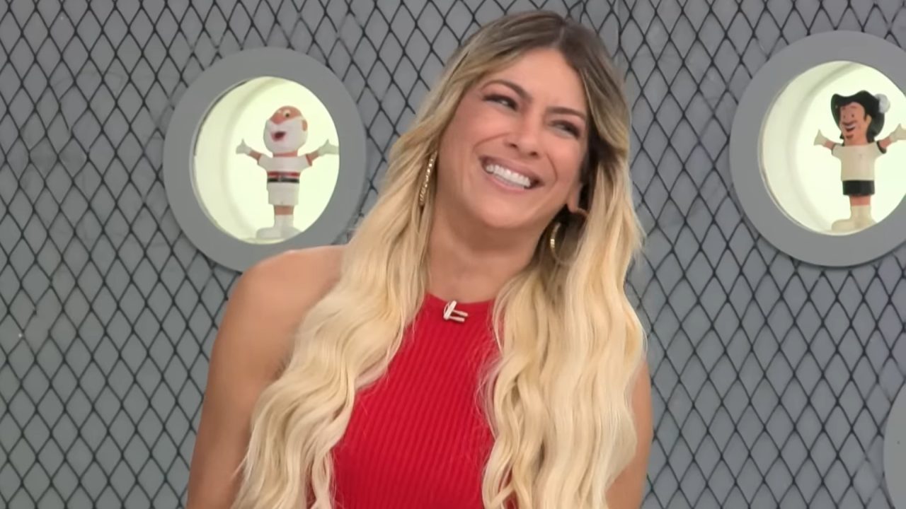 A imagem mostra Renata Fan, uma mulher sorridente com cabelos longos e loiros, vestindo um vestido vermelho sem mangas. Ela está em um estúdio com fundo cinza e detalhes em forma de rede. Atrás dela, há duas pequenas janelas circulares com bonecos de times de futebol exibidos dentro delas. O clima é leve e descontraído, no programa Jogo Aberto, da Band